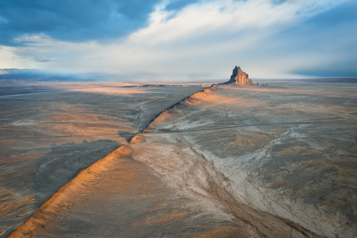 Aerial view of Shiprock, New Mexico, with a sunlit volcanic ridge across the desert beneath a blue and cloud-filled sky